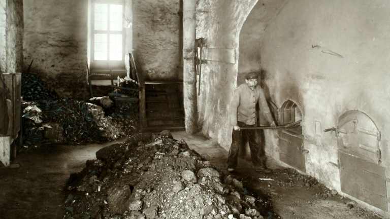 Historisches Foto: Blick in einen Heizungsraum, im Vordergrund Kohlehäufen, rechts ein Arbeiter an gemauerten Öfen