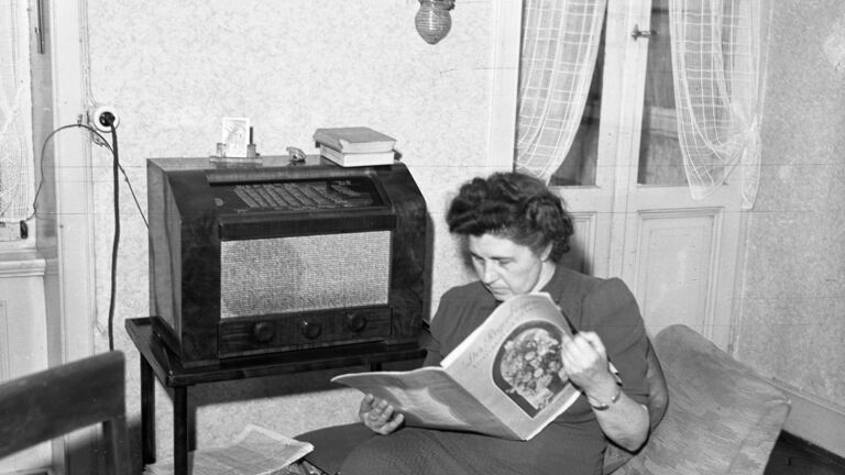 schwarz-weiß Foto einer Frau die auf eine Sessel sitzt, Zeitung liest und nebenher Radio hört