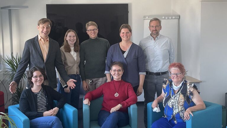 Eine Gruppe von Menschen, fünf stehend und drei in Sesseln sitzend lachen in die Kamera