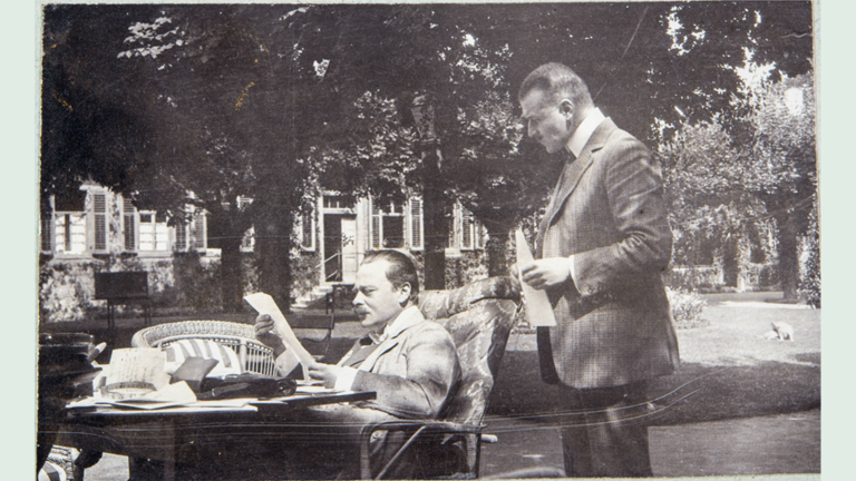 Historisches Foto: Blick in einen Garten, im Vordergrund ein Tisch, an dem ein lesender Mann sitzt, neben ihm ein stehender Mann