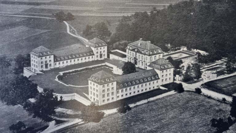 Schwarz-Weiß-Luftbild eines großen, symmetrischen Gebäudekomplexes mit mehreren Flügeln und Innenhof, umgeben von Wegen, Feldern und dichtem Wald