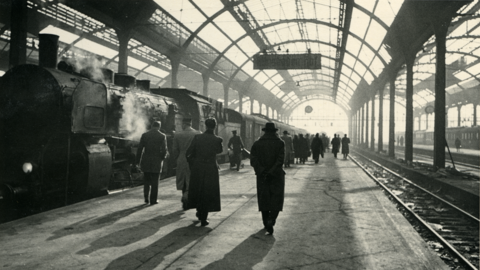Historisches Foto aus dem Hauptbahnhof Wiesbaden um 1955: Links wartet ein von einer Dampflok gezogener Personenzug, auf dem Bahnsteig in der Mitte halten sich Reisende auf (HHStAW, 3008/2, 30155)