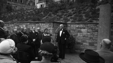 Historisches Foto: Redner am Mikrofon, zuhörende Menschen, rechts eine Steinstele