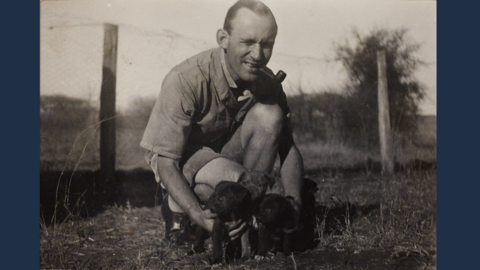 Wilhelm Wilbrand in Kenia (HStAD, O 13, 679) Historisches Foto: Ein Mann hockt auf dem Boden, vor sich drei Hundewelpen