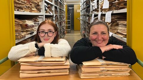Fami-Auszubildende im Hessischen Landesarchiv Modernes Foto: Zwei Frauen stützen sich auf Aktenstapel und blicken den Betrachter lächelnd an.