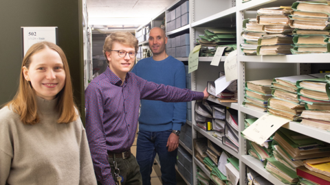 Das Team der Übernahme aus dem Hessischen Hauptstaatsarchiv Wiesbaden. Modernes Foto: Links eine junge Frau, dann ein Mann, im Hintergrund ein weiterer Mann, rechts ordnergefüllte Regale