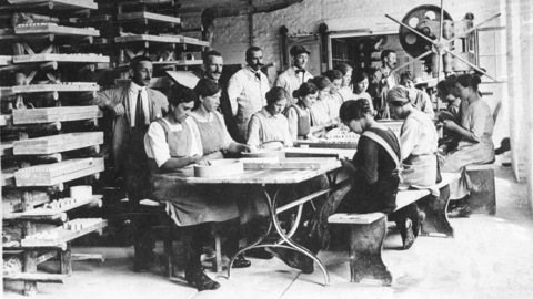Moeller AG Werk (Lochmühle) bei Schlangenbad, um 1900 (HHStAW, 3008/2, 22000) Historisches Foto: Frauen arbeiten an einem Tisch in einer Werkshalle, Männer im Hintergrund sehen zu