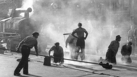 Asphaltierungsarbeiten in der Rheinstraße in Wiesbaden, 1956 (HHStAW, 3008/2, 7537) Historisches Foto: Männer in Dunstschwaden gießen Teer, dahinter die Teerkochmaschine