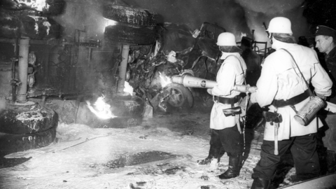Löscharbeiten nach einem LKW-Unfall, um 1955 (HHStAW, 3008/47, 3639) Historisches Foto: Links ein umgestürzter brennender LKW, rechts zwei Feuerwehrleute beim Löschen und ein Polizist