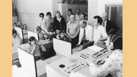Modernes Foto: Jugendliche mit Kopfhörern sitzen an Pulten, rechts eine Besuchergruppe, vorne rechts ein Lehrer an einem Schaltpult