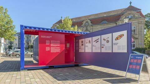 Blick in die Wanderausstellung #StolenMemory Modernes Foto: ein geöffneter blau-roter Schiffscontainer auf einem Platz