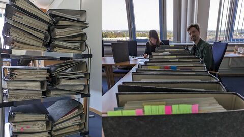 Aktenstapel und zwei Personen an einem Tisch mit Akten