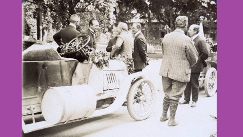 Historisches Foto: Links ein blumengeschmückter Rennwagen, daneben eine Gruppe von Herren im Gespräch