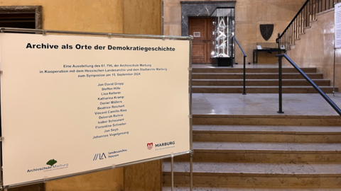 Tafel mit der Aufschrift „Archive als Orte der Demokratiegeschichte“, darunter die Namen der an der Ausstellung beteiligten Personen. Im Hintergrund der Treppenaufgang und das Foyer des Hessischen Staatsarchivs Marburg