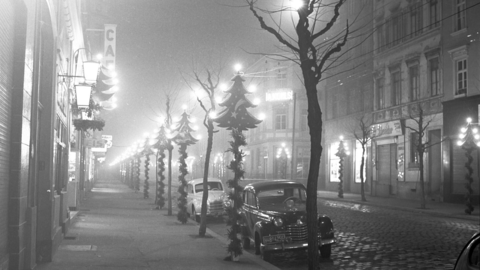 Historisches Foto: Blick in eine weihnachtlich dekorierte Straße ohne Passanten, am Straßenrand einige parkende Autos