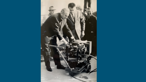 Foto: Ein Herr im Anzug betätigt eine Pumpe, dahinter weitere Männer