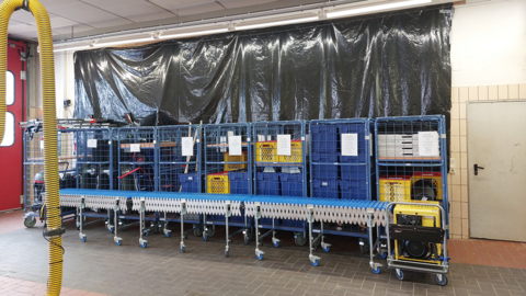 Foto: Farbige Boxen in Gitterboxen nebeneinander aufgereiht. Dahinter eine Wand mit schwarzer Folie