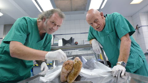 Zwei Männer in grünem Arbeitskittel stehen sich an einem Sektionstisch gegenüber. Auf dem Tisch liegt mit Plastik umhüllt eine Puppe, die eine menschliche Leiche darstellen soll. Zu sehen sind aber nur die schmutzigen Füße 