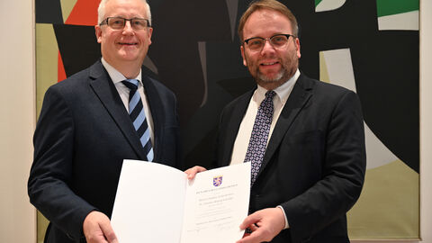 Minister Timon Gremmels überreicht Dr. Kistenich-Zerfaß die Ernennungsurkunde zum Präsidenten des Hessischen Landesarchivs Zwei Männer im Anzug halten gemeinsam eine Urkunde