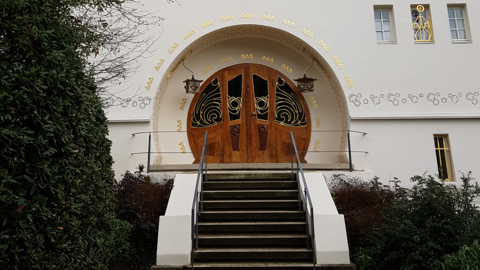 Spiel mit Materialien, Farben und Formen: Treppe und Portal des "Großen Glückerthauses" auf der Mathildenhöhe, 2025 Foto: Treppenaufgang zu einem Haus mit auffällig rundem Eingangsportal aus verschiedenen Hölzern und filigranen Metallverzierungen