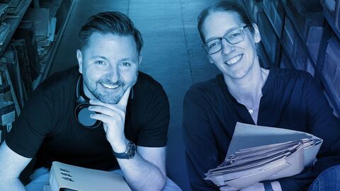 Das Cover des Podcast, moderiert von Jan-Hendrik Evers und Maria Kobold Ein Mann und eine Frau sitzen im Schneidersitz auf dem Boden und halten Akten in der Hand. Das Bild ist bläulich eingefärbt. Zu sehen ist in der unteren Hälfte in weißer Schrift auf blauem Grund in einer Box: Aufgeschlossen - Türen zur Geschichte