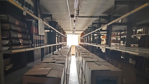 Verpackte Dienstbibliothek des Staatsarchivs Marburg Blick zwischen die Regale eines Magazins, in den Fächern liegen Bücher, im Gang stehen zwei Reihen Kartons