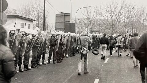 Polizisten schützen das Gelände der Firma NUKEM in Hanau-Wolfgang bei einer Demonstration gegen die dortige Produktion von Brennelementen am 8. November 1986 (HHStAW, 3008/1, 79243). Aufnahme in schwarz-weiß von Polizisten, die mit Schutzkleidung und Schildern eine Straße säumen, auf der Menschen laufen