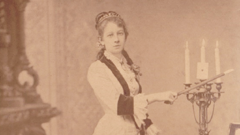 Die Zauberkünstlerin Emma Stengel (HHStAW, 1142, 12) Historisches Foto: Porträt einer Frau, die in der rechten Hand einen Zauberstab hält, daneben ein Kerzenleuchter mit drei brennenden Kerzen und einer Spielkarte