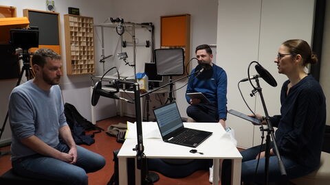 Zusammen mit Dr. Jochen Lehnhardt bei der Aufnahme der Folge im Tonstudio der Philipps-Universität Marburg zwei Männer und eine Frau sitzen an einem Tisch, vor ihnen Mikrofone