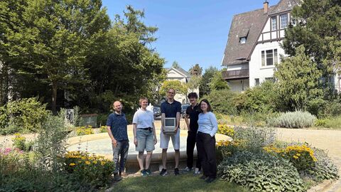 Die Auszubildenden zusammen mit Dr. Christoph Schmidt (Mitte), Leiter der Digitalen Dienste im Hessischen Landesarchiv Abgebildet sind fünf Personen, die in einem grünen mit Blumen bepflanzten Park vor einem Brunnen stehen. In der Mitte steht der Ausbilder neben ihm jeweils zwei junge Frauen und zwei junge Männer. Alle sind in einer Reihe aufgestellt und lächeln in die Kamera, der Ausbilder hält einen alten PC in der Hand.