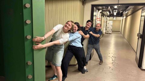 Ausbildung öffnet Türen...auch wenn es manchmal schwer sein kann! Auf dem Bild ist ein langer gelber Gang zu sehen, eine schwere grüne Tresortür aus Stahl wird von vier Personen aufgezogen. Es sind zwei junge Frauen und zwei junge Männer, die sich hintereinander als Kette aufgestellt haben und lachend die Tür aufziehen.