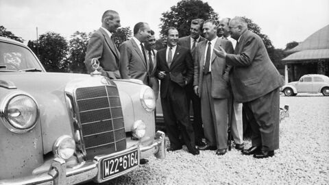 Historisches Foto: Vorne links die Front eines Mercedes, dahinter eine Gruppe von acht Männern in Anzügen