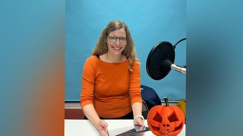 Nicht ganz unpassend zu Halloween sprechen wir mit Dr. Constanze Sieger über die historische Debatte zum Thema "Scheintod" eine Frau mit orangenem Oberteil sitzt an einem Tisch, in der Hand ein Tablett, vor ihr ein Mikro und rechts neben ihr ein Kürbis