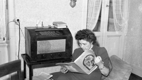 Beliebtes Medium in der ersten Hälfte des 20. Jahrhunderts: Das Radio schwarz-weiß Foto einer Frau die auf eine Sessel sitzt, Zeitung liest und nebenher Radio hört