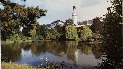 Idyllischer Blick über den Weiher im Schlosspark auf das Schloss Bad Homburg. Ablichtung für das Kalenderblatt Juli 1986 des Hessenkalenders (HStAD Best. R 4, Nr. 14131 UF) Fotografie eines mit Bäumen umrandeten Sees, im Hintergrund ein Schloss mit einem hohen Turm