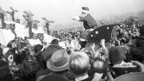 Historisches Foto: Ein als Weihnachtsmann verkleideter Mann, der in einem von Papp-Rentieren gezogenen Schlitten sitzt, wirft Süßigkeiten in eine Menschenmenge
