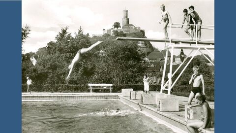 Historisches Foto: Kinder springen von einem Zwei-Meterbrett in ein Schwimmbecken, im Hintergrund auf einem Hügel eine Burgruine