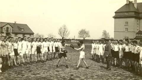 Historisches Foto: Links und rechts eine Reihe von Männern, in der Mitte zwei boxende Männer