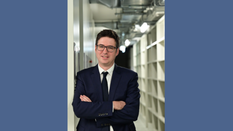 Mann in Anzug mit Brille und verschränkten Armen steht in einem Gang mit Regalen