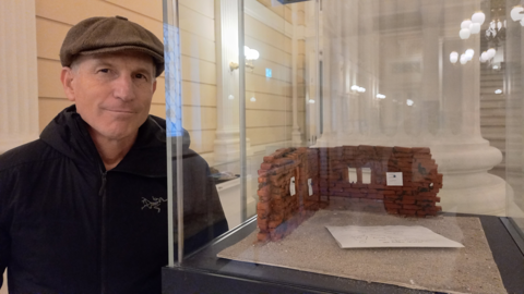 Foto eines Mannes mit Mütze neben einer Vitrine, in der sich eine Mauerecke aus Ziegelsteinen befindet