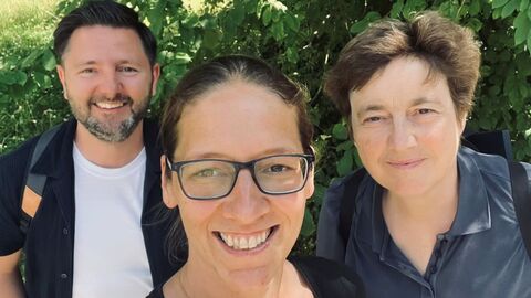 Drei Personen lächeln in de Kamera bei einem Selfie im Freien; im Hintergrund sind grüne Pflanzen zu sehen