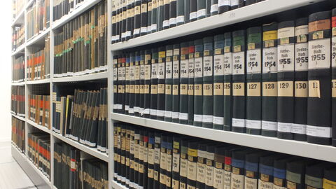 Archivmagazin mit langen Regalreihen, gefüllt mit gebundenen Amts-und Registerbänden (u.a. "Sterbebuch") aus den 1950er Jahren, beschriftet mit Jahreszahlen und Signaturen.