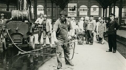 Historisches Foto: in der Bildmitte ein Arbeiter mit einem Wasserschlauch mit Sprühdüse, links Arbeiter an einer Pumpmaschine, links ein Bahnbeamter, im Hintergrund zuschauende Passagiere