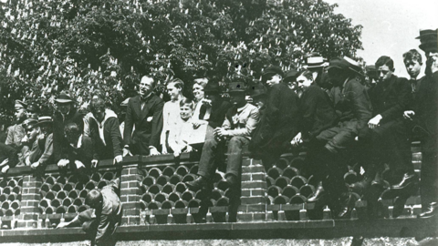 Historisches Foto: Männer und Jungen sitzen auf einem Ziegelzaun bzw. stehen dahinter