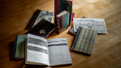 Modernes Foto: Auf einer Holzfläche liegen Büchlein, eines davon aufgeschlagen. Der handschriftliche Text ist teilweise geschwärzt.